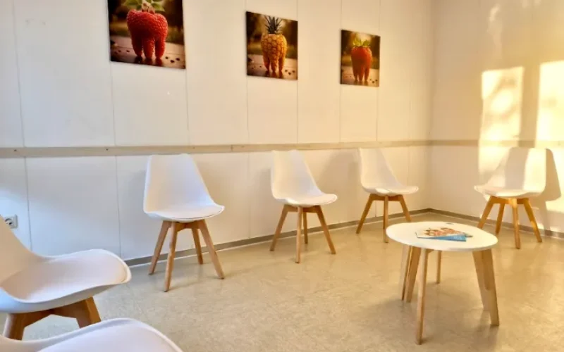 Ein heller Warteraum mit fünf weißen Stühlen, einem runden Tisch und Bildern von Obst an der Wand.